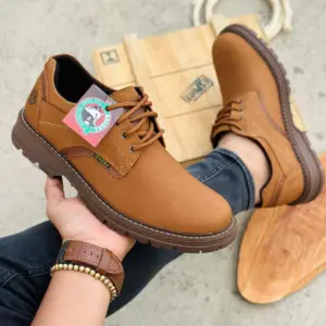 Zapato sport de cuero genuino color marrón claro con suela track y cordones, sostenido en la mano con etiqueta visible sobre fondo rústico.